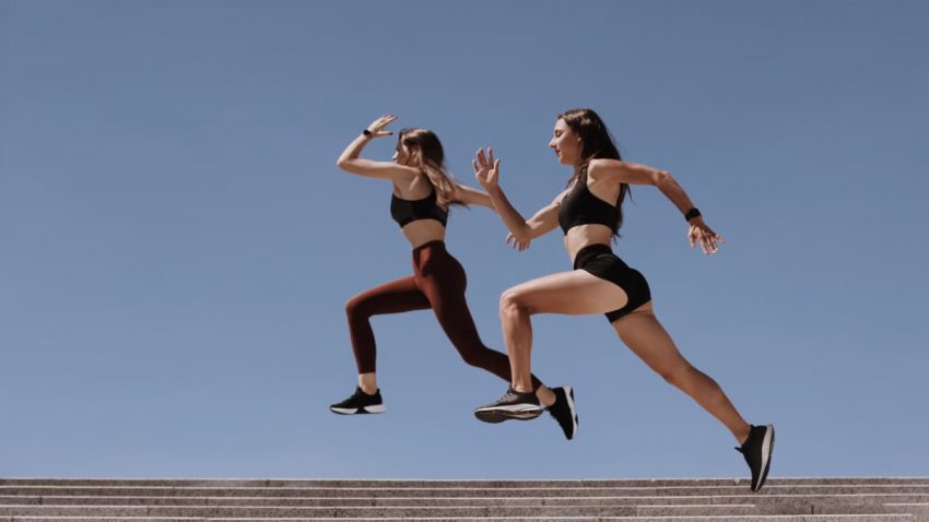 Two women in active wear leap energetically on stairs against a clear blue sky.